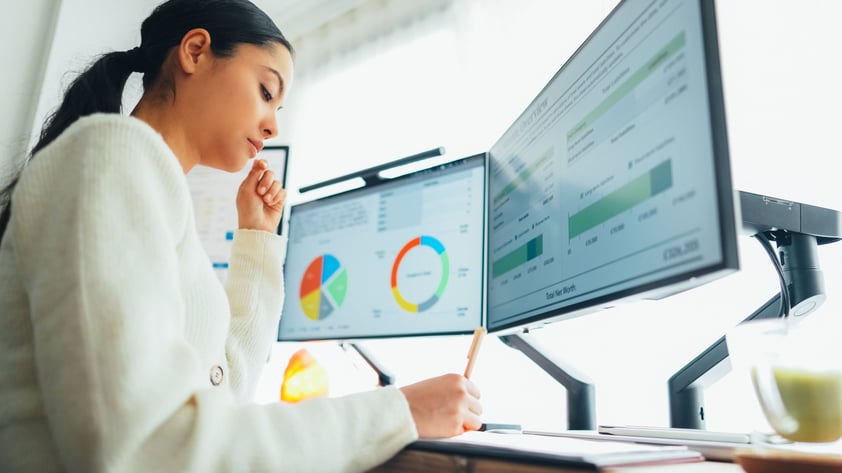 woman writing on desk with two screens of charts and graphs visible